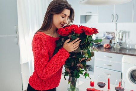 Young woman found bouquet of roses, wine and gift box on kitchen. Happy girl hugging flowers. Valentines day surpriseの写真素材