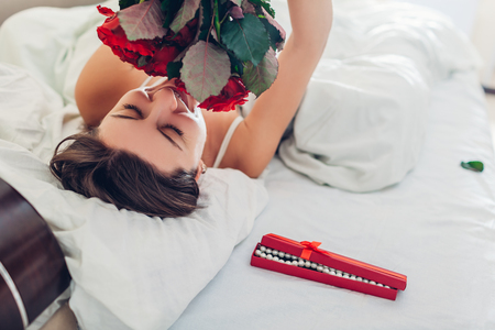 Young woman found bouquet of roses with jewellery in gift box in bed. Happy girl smelling flowers. Women's day surpriseの写真素材