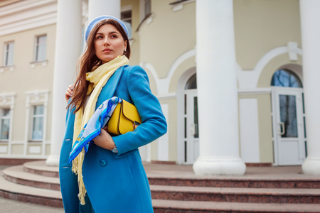 Young woman in trendy blue coat walking on city street holding stylish handbag. Spring female clothes and accessories. Fashionの写真素材