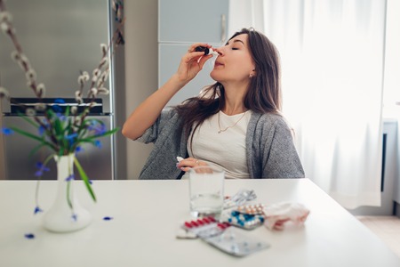 Spring allergy. Tired woman using nasal drops against seasonal allergy and taking pills on kitchen at home. Healthcare and medicineの写真素材