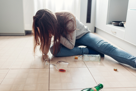 Female alcohol addiction. Young woman woke up on kitchen floor after party surrounded with wine bottles and having hangoverの写真素材