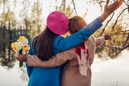Happy senior mother with flowers and her adult daughter hugging by spring river at sunset. Mother's day concept. Family valuesの写真素材