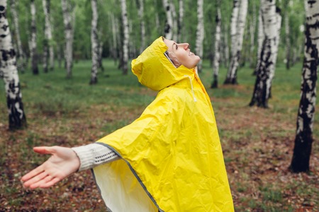 Happy young woman in yellow raincoat walking in spring forest under rain and having fun raising arms. Enjoying lifeの写真素材