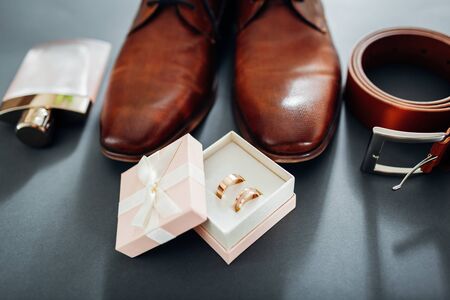Groom's wedding day accessories. Brown leather shoes, belt, perfume, golden rings in box. Male fashionの写真素材