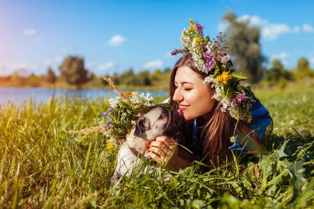 Pug dog and its master chilling by lake wearing flower wreaths. Happy puppy licking woman in summer parkの写真素材