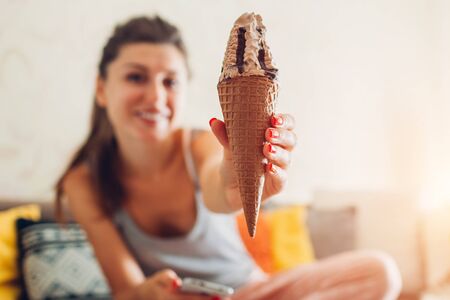 Young woman eating chocolate ice-cream in cone sitting on couch at home. Girl holding and showing dessertの写真素材