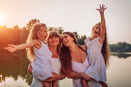 Family walking by summer river at sunset. Mother and grandmother holding kids and laughing. Three...