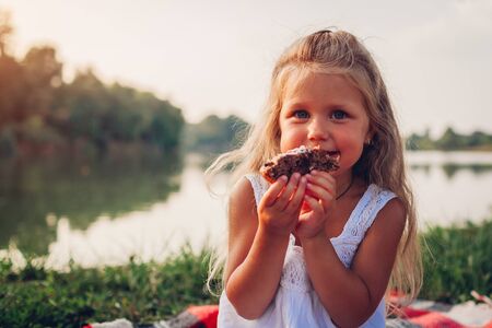 Little girl eating cake on family picnic by summer river. Child holding piece of pie and smilingの写真素材