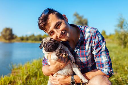Man hugging pug dog in park. Happy man walking with pet by summer lakeの写真素材