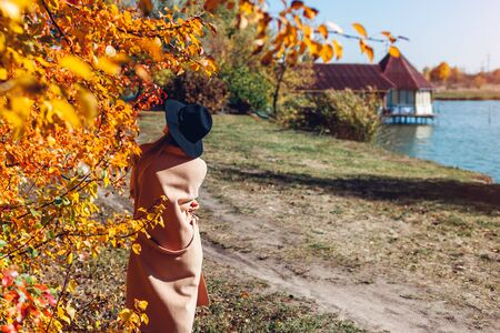 Autumn landscape. Young woman walking in autumn forest by lake. Stylish girl wearing hat and coatの写真素材