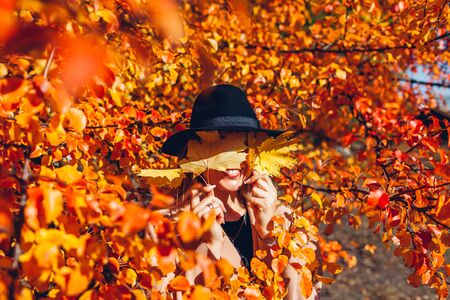 Autumn fun. Young woman walking in autumn forest covering eyes with leaves. Girl having funの写真素材