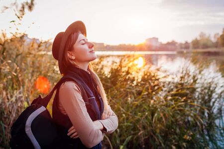 Traveler with backpack relaxing by autumn river at sunset. Young woman breathing deep feeling happy and free. Active lifestyleの写真素材