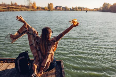 Traveler with backpack relaxing by autumn river at sunset. Young woman raised arms feeling free and happy sitting on pier. Active lifestyleの写真素材