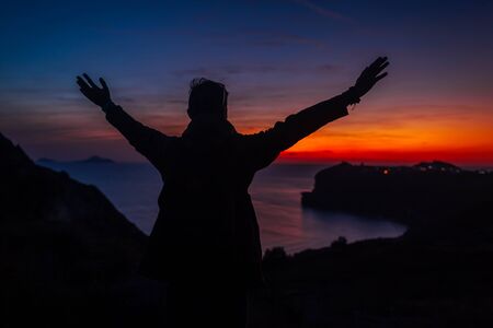 Man traveler silhouette hiking at sunset on Santorini island. Happy tourist raised arms feeling...