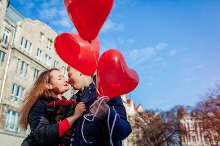 Valentines day date. Man and woman about to kiss outdoors. Couple walking with roses flowers and heart shaped balloons. Womens day surprise giftの写真素材