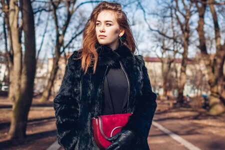 Female fashion. Stylish woman wearing fur coat and holding red purse. Spring clothes accessories. Outdoors portrait on city streetの写真素材