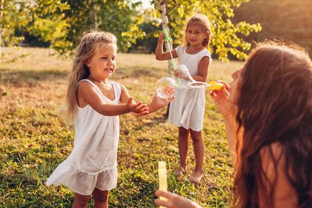 Mother helps daughters to blow bubbles in summer park. Kids having fun playing and catching bubbles outdoorsの写真素材