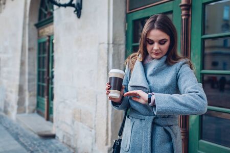 Young stylish woman looking at hand watch drinking coffee on city street waiting for friends. Spring female fashionの写真素材