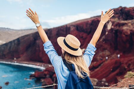 Tourist looking at Red beach landscape from view point in Akrotiri Santorini Greece. Happy woman traveler with backpack raising armsの写真素材