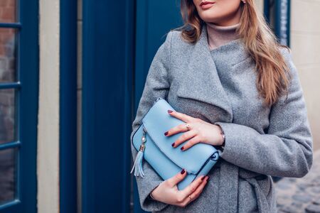 Female purse. Fashionable spring accessories. Stylish woman wearing grey coat and holding blue handbag on street by cafe window outdoors. Close upの写真素材