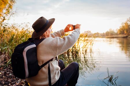 Tourist with backpack taking photos using smartphone of river at sunset. Woman travels admiring spring natural landscape. Exploring the worldの写真素材