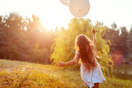 Happy little girl running with baloons in hand. Kid having fun in summer park at sunset. Outdoors activitiesの写真素材