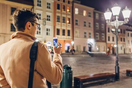 Tourist man taking photos on smartphone at night on Market square in Krakow Poland. traveler walking...