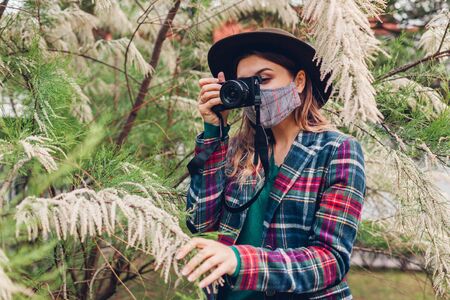 Woman photographer taking photos using camera in summer garden wearing coronavirus mask.