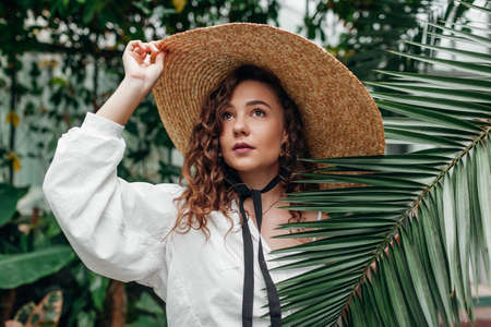 Stylish summer straw sun hat. Portrait of young fashionable woman wearing retro accessories in tropical exotic garden.の写真素材