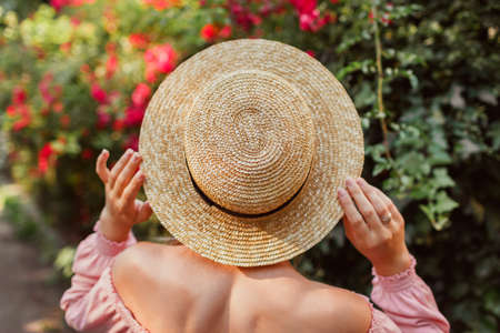 Summer straw sun hat. Girl walking by blooming roses in garden. Fashionable accessories. Back view