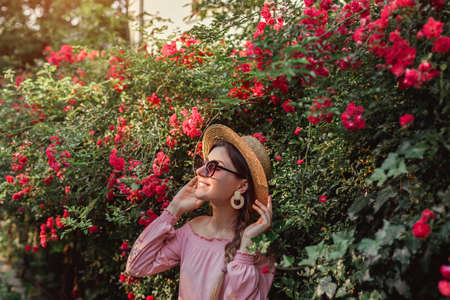 Outdoor portrait of woman walks by blooming roses in summer garden. Stylish happy girl enjoys flowers in park. Fashion outfit.の写真素材