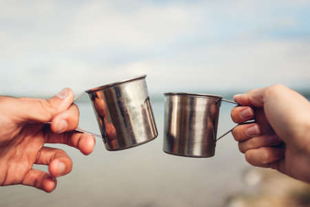 Couple of travelers holding hot tea in metal cups clinking. people drink warm beverages by lakeの写真素材