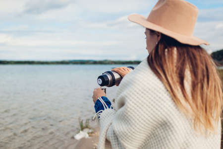 Young girl tourist pours hot tea out of flask by autumn lake enjoying landscape. Woman in hat...