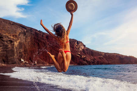 Sexy woman in bikini walks on Red beach in Santorini by sea. Girl jumps raising arms feeling happy and free having fun enjoying summer vacationの写真素材