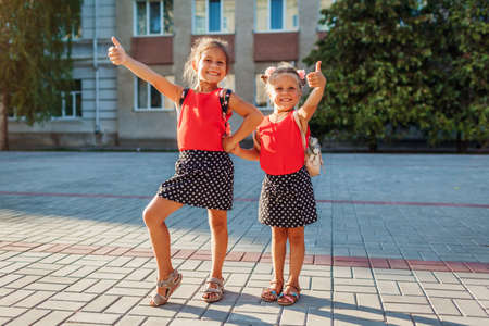 Happy sisters girls wearing backpacks and showing thumbs up. Kids pupils having fun outdoors school building. Education. Back to schoolの写真素材