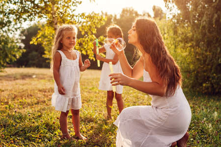 Mother and daughters blow soap bubbles in summer park. Kids having fun playing, catching bubbles having fun outdoors. Family spending time togetherの写真素材