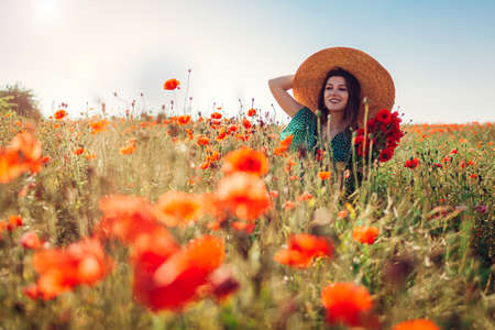 Young woman picking poppies flowers walking in summer field enjoying landscape. Stylish happy girl wearing straw hatの写真素材