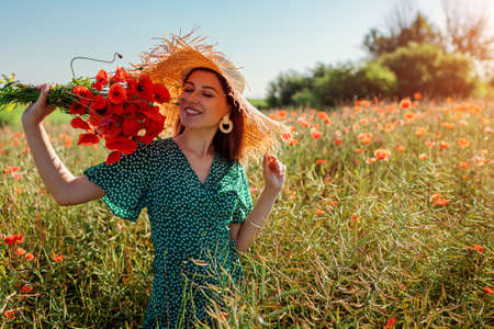 Young happy woman smelling bouquet of poppies flowers walking in summer field. Stylish girl wearing straw hatの写真素材