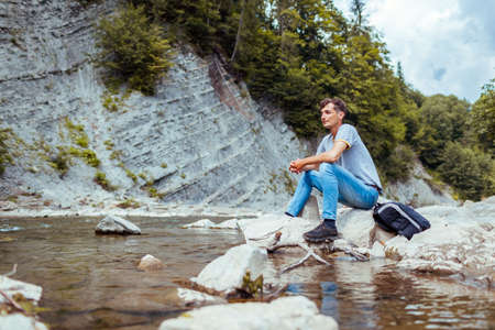 Young man tourist relaxing by mountain river enjoying natural landscape. Traveller backpacker sitting on rock. Summer tripの写真素材