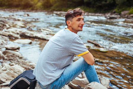 Tourist man relaxing by mountain river enjoying wild natural landscape. Traveler backpacker sitting on rock. Summer tripの写真素材