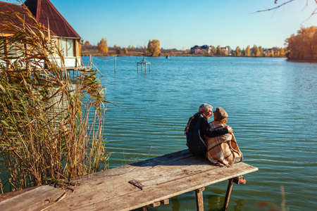Senior family couple relaxing by autumn lake. Happy man and woman enjoying natural landscape and hugging sitting on wooden pierの写真素材