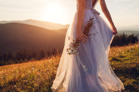 Bride wearing blue wedding dress holding bouquet in mountains at sunset. Woman walking on meadow in flowersの写真素材