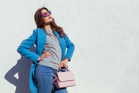 Young woman holding stylish purse and wearing trendy blue coat, sunglasses. Autumn female clothes and accessories. Fashionの写真素材