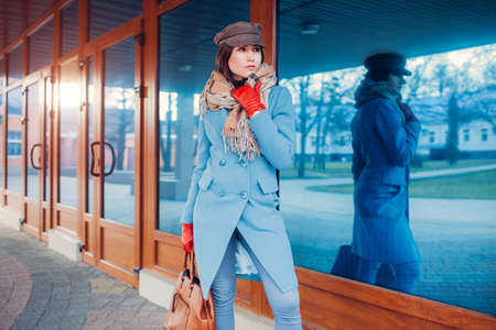 Portrait of woman wearing blue coat, cap and gloves holding handbag outdoors. Autumn female clothes and accessories. Fashionの写真素材