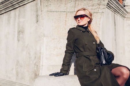 Portrait of stylish young woman in coat sunglasses with purse outdoors. Autumn fashion female clothes accessories. Trendy modern outfitの写真素材
