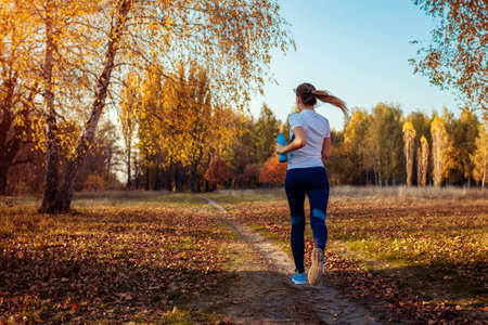 Runner training in autumn park. Woman running with water bottle at sunset. Active healthy lifestyle. Workout. Fall activities. Sportの写真素材