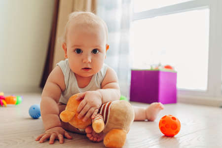 Little newborn baby playing with toys sitting on floor at home. Kid looking at camera holding teddy bearの写真素材