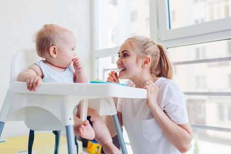 Mother feeding little baby with fruits at home. Family eating together. Infant sitting in chair for...