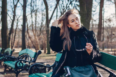 Portrait of stylish woman wearing fur coat holding sunglasses handbag. Autumn clothes and accessories. Girl relaxing on bench in parkの写真素材