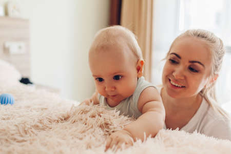 Mother playing with her newborn baby son at home. Family having fun. Curious infant touching blanket on bedの写真素材